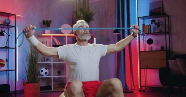 Домашнє тренування, де добре виглядає активний спортивний старий бородатий спортсмен у тренувальному одязі, сидячи на фітболі і роблячи вправи з розширювачем у вечірній час — стокове відео