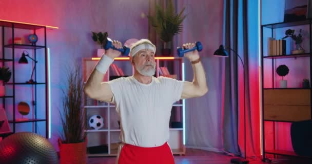 Retrato de atleta de 70 años de edad barbudo deportivo activo atractivo en ropa deportiva que hace ejercicios con pesas durante el entrenamiento de la noche en casa — Vídeos de Stock