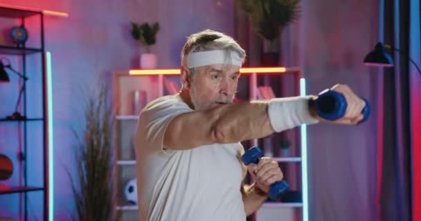 Domácí večerní cvičení, kde atraktivní aktivní aktivní soustředěný šedovousý starší muž ve sportovním oblečení dělá boxerské cvičení s činkami, zblízka — Stock video