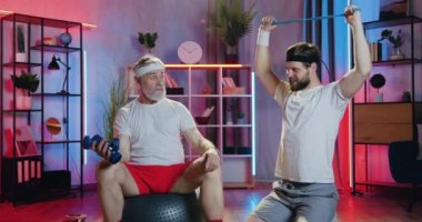 Yakışıklı, sportif, son sınıf öğrencisinin portresi. Spor yaparken, sakallı, pozitif torunu direnç bandını esnetirken halterin üzerinde oturan dambıllarla egzersiz yapıyor. Sonra da beşlik çakıyorlar.