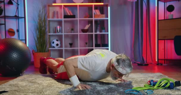 Goed uitziende sterke sportieve bebaarde oude man in sportkleding die 's avonds push-ups vanuit de vloer thuis doet, gezond lifestyle concept — Stockvideo