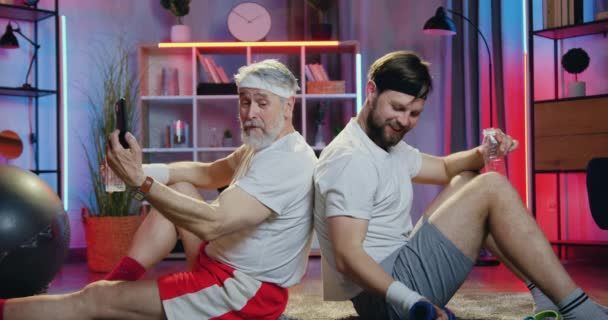 Good-looking smiling active sporty bearded different ages athletes sitting on the floor back to back,one of them using smartphone,another doing exercises with dumbbells — Stock Video