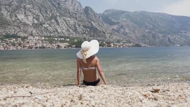 Rückansicht einer Frau mit Strohhut, die am felsigen Strand vor dem Meer sitzt und das gegenüberliegende Schiff begrüßt. Sommerferien — Stockvideo