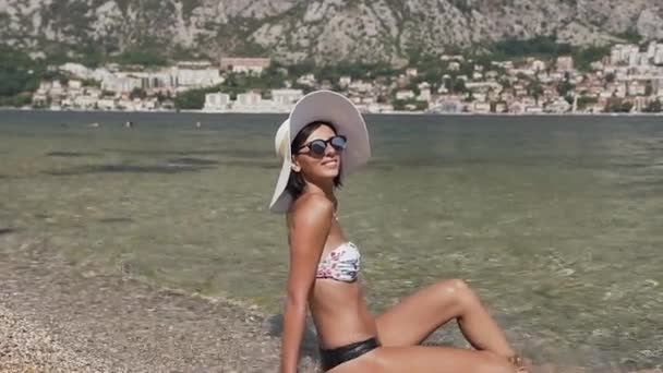 Uśmiechnięta szczupła kobieta w bikini, szeroki kapelusz i okulary przeciwsłoneczne na skalistej plaży w pobliżu morza w wakacje lata. Na zewnątrz — Wideo stockowe