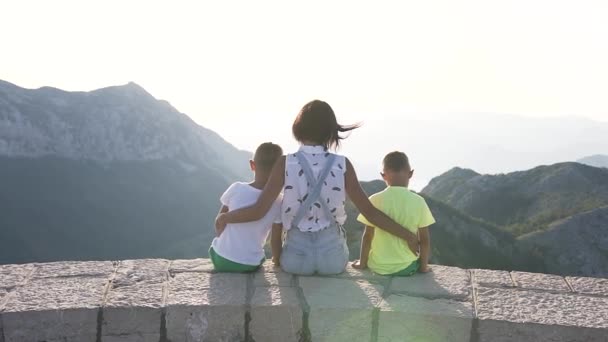 Задний вид молодой матери, обнимающей двух детей, идущих по вершине горы на открытом воздухе, глядя на красивый пейзаж на закате — стоковое видео