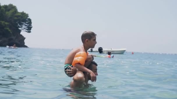 Portrait shot of little boy who does jump somersault off his fathers shoulder in the beautiful blue sea during summer holiday — Stock Video