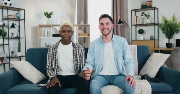 Schön lächelnd glücklich liebend multirassisch männlich homosexuell pärchen holding by ihre hände und looking at camera sitting on the couch im gemütlichen zimmer — Stockvideo