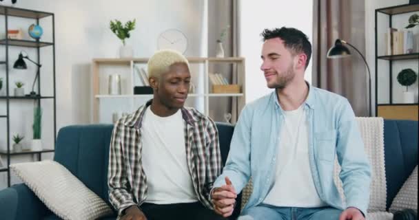 Glimlachen gelukkig amoureuze multiraciale gay partners op zoek met tederheid in de ogen van elkaar en houden door de handen in de voorkant van de camera thuis, close-up — Stockvideo