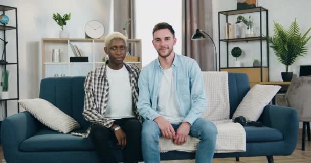 Likable gelukkig jong Afrikaans amerikaans gay knuffelen zijn europees vriendje door de schouder terwijl ze beiden zitten op de bank thuis en kijken naar camera met glimlach — Stockvideo