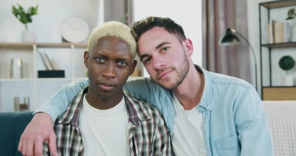Detailní záběr dobře vypadající spokojený usmívající se mladý multiiraciální gay pár, který sedí před kamerou se skloněnými hlavami a jeden z nich položí ruku na rameno svých partnerů — Stock video