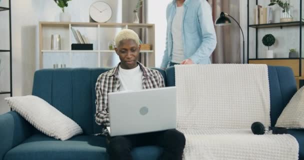 Attraktivt positivt lugn afrikansk amerikansk gay sitter på soffan hemma och använder laptop när en annan europeisk partner passar bakom soffan och ömt kramar honom — Stockvideo