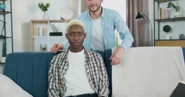 Kanepenin arkasında dikilen ve kanepede oturan Afro-Amerikan ortağına sarılan mutlu ve mutlu bir genç gay. İkisi de oturma odasında kameraya bakıyor.
