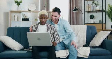 LGBT toplum konsepti. Yakışıklı, gülümseyen, melez, eşcinsel bir çiftin oturma odasındaki kanepede oturup dizüstü bilgisayardaki video çağrısının keyfini çıkardığı yer.