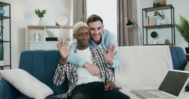 Porträtt av sympatisk glad kärleksfull ung blandad ras homosexuella par som tittar in i kameran med glada ansikten, en av dem kramar en annan vid halsen medan han viftar med händerna i kameran — Stockvideo