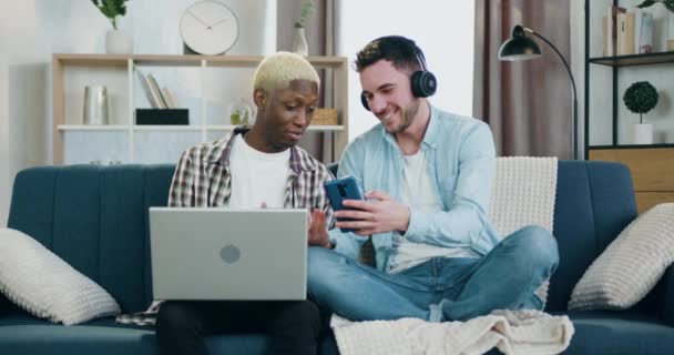 Homosexual relationship concept where loving smiling happy young gay couple spending their joint leisure at home,one of them using laptop while another listening music in headphones — ストック動画