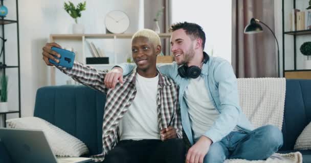 행복 한 미소를 짓고 있는 젊은 동성 커플 이 집 소파에 앉아 셀카와 전화 통화를 하고 동성애 관계 개념을 만들고 있습니다. — 비디오