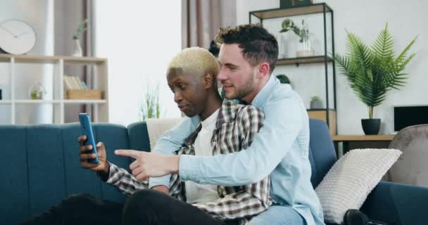 Zblízka roztomilé šťastný úsměv multiraciální mladý homosexuální pár v lásce, která relaxuje na gauči doma ve dne a použití smartphone, lidé s nekonvenční sexuální orientace vztahu — Stock video