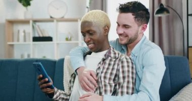 Yakışıklı, mutlu, tutkulu, tutkulu bir eşcinsel. Siyah tenli erkek arkadaşını kucaklıyor. Evde kanepede oturup akıllı telefon kullanırken.