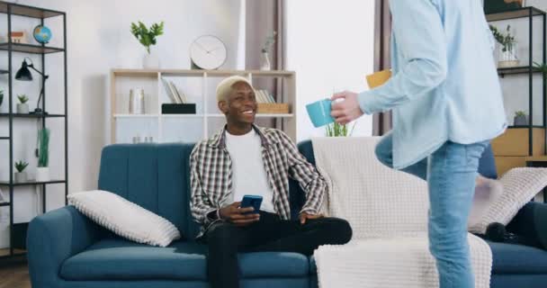 Pozitivní uvolněný černošky-skinned gay s blond vlasy sedí na gauči doma a používá svůj mobil, když usměvavý muž partner přináší šálky čaje a sedí vedle něj — Stock video