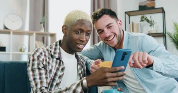 Ritratto di carino amorevole felice sorridente razza mista coppia gay che seduti insieme sul divano in soggiorno e utilizzando telefono trascorrere il tempo libero comune — Video Stock