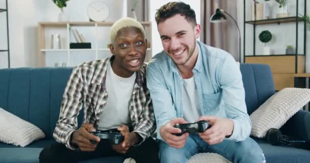 Close up de atraente feliz sorrindo brincalhão misto raça jovem gay casal no amor que sentado no sofá em casa e jogar videogames comemorando a vitória com alta cinco — Vídeo de Stock