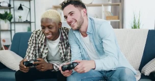 Attrayant heureux souriant amour mixte race gay couple profiter de leurs loisirs à la maison à jouer à des jeux sur le téléviseur en utilisant joysticks, homosexuel relation concept — Video
