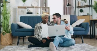 Şirin, mutlu, gülümseyen, melez eşcinsel bir çiftin portresi rahat bir odada koltuğun yanında oturuyor ve dizüstü bilgisayar kullanıyor, Afrikalı Amerikalı ortak Avrupalı erkek arkadaşlara elini sürüyor.