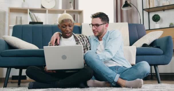Närbild på godmodigt leende nöjda unga blandras homosexuella par som tillbringar gemensam fritid hemma och använda laptop sitter på golvet nära soffan — Stockvideo