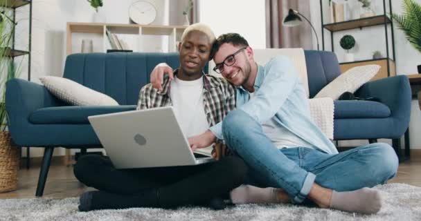 Concepto de relatioship homosexual donde dulce feliz sonriente despreocupado amoroso mestizo pareja masculina descansando juntos en el suelo en casa y el uso de la computadora portátil — Vídeos de Stock