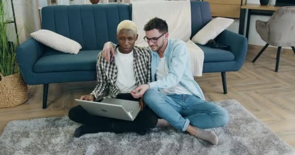 Concepto LGBT-comunidad donde lindo feliz positivo joven pareja gay multiétnica sentado en el suelo con abrazos y la aplicación de la computadora simultanepusly hablando entre sí — Vídeos de Stock