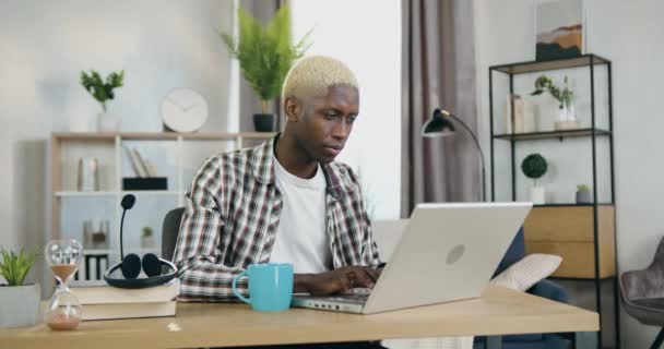 Chico joven concentrado y bien parecido de piel negra con el pelo rubio sentado en su lugar de trabajo en la oficina en casa y trabajando en el portátil — Vídeos de Stock