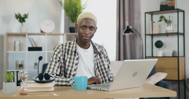 Portret van goedgehumeurd glimlachen gelukkig jong zwart-gevild gay met blond haar die poseren op camera op zijn werkplek thuis en tonen symbool ok — Stockvideo