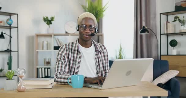 Retrato de adorável satisfeito sorrindo jovem preto de pele gay com cabelo loiro no fone de ouvido que sentado na frente da câmera no escritório em casa durante o trabalho no laptop, conceito LGBT-comunidade — Vídeo de Stock