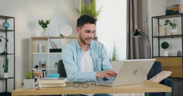 Snygg nöjd leende ung man sitter vid bordet i mysiga vardagsrum och chatta med vänner på datorn, framifrån, slow motion — Stockvideo