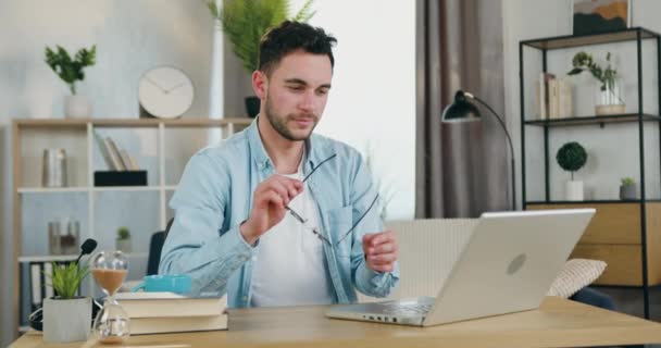 Snygg koncentrerad ung smart kille sätta på sina glasögon och börjar arbeta på datorn sitter på arbetsplatsen i hemmakontoret — Stockvideo