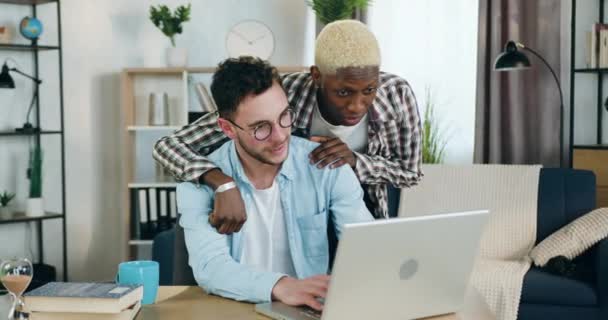 Homosexual relationship concept where cute smiling satisfied black-skinned guy with same sex orientation standing and putting his hand on positive joyful partners shoulder which working on laptop at — ストック動画