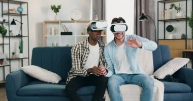 LGBT toplumu ve modern teknoloji kavramı. Evdeki koltukta oturup el ele tutuşan ve sanal gerçeklik kulaklığı takan sevimli, mutlu, genç erkek partnerler.