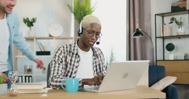 Mignon heureux souriant jeune gay à la peau noire dans les écouteurs travaillant sur ordinateur portable dans le bureau à la maison lorsque gai aimant partenaire européen approchant de lui et étreignant par le cou — Video