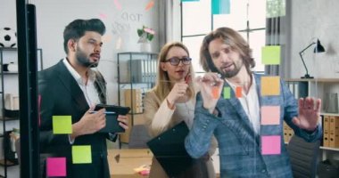 İyi görünümlü başarılı takım modern ofiste cam duvarın yanında duruyor ve yapışkan notlar, iş konsepti kullanarak yeni fikirler üretiyorlar.