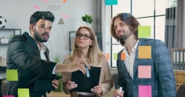 Yapışkan notlar üzerinde çizimler ve hatırlatıcılar kullanarak cam duvarın yanında yeni bir başlangıç projesini tartışan olumlu erkek ve kadın yaratıcıların ön görünümü