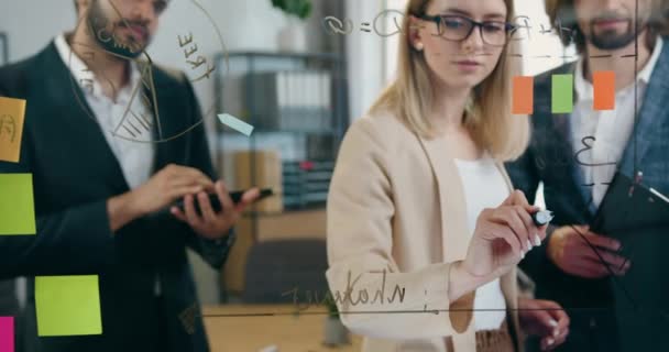 Atractiva mujer inventiva dibujando gráficos en la pared de vidrio para sus colegas masculinos durante el trabajo con ellos sobre el nuevo proyecto de puesta en marcha en la oficina moderna, vista frontal — Vídeos de Stock