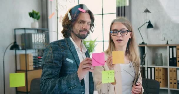 Atractivo hombre y mujer experimentados creativos seguros de pie detrás de la pared de vidrio en la habitación de la oficina e ideas de lluvia de ideas para el proyecto conjunto utilizando notas adhesivas — Vídeos de Stock