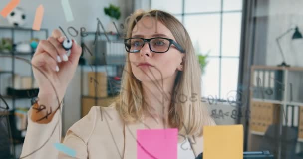 Primer plano de la mujer rubia creativa y atractiva en gafas que escribe nuevas ideas en tablero de vidrio trabajando sobre el proyecto de puesta en marcha en la sala de oficina — Vídeos de Stock