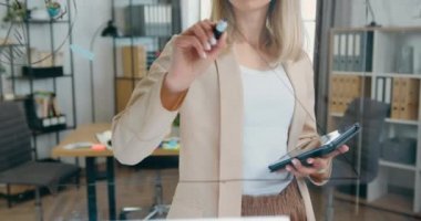 Ofis odasındaki cam duvarın yanında duran, grafik üzerine ok çizen ve bu duvardan kameraya bakan yüksek yetenekli sarışın kadın-şirket çalışanı.