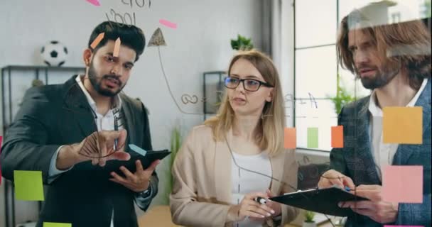 Atractivo equipo creativo joven hábil trabajando juntos durante el proyecto de puesta en marcha y la grabación de ideas en la pared de vidrio en la oficina moderna, concepto de estrategia de la empresa — Vídeo de stock