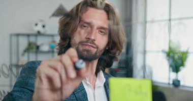 Modern ofis odasındaki cam tahtaya notlar yazan iyi görünümlü, kendine güveni yüksek, başarılı, sakallı, ofis çalışanı.