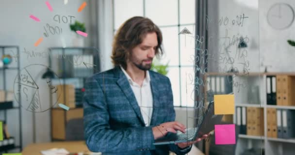 Camera shoot on likable positive hardworking experienced bearded worker which typing on laptop near glass board in modern office room,workflow concept — Stock Video