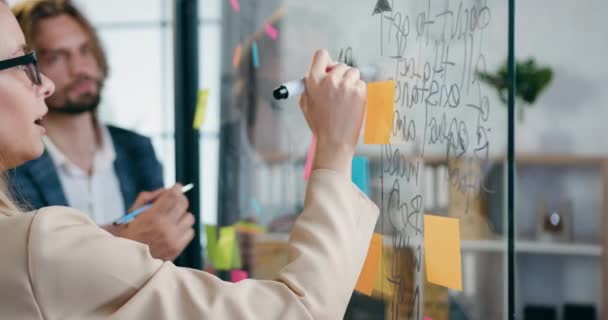 Likable confident successful professional male and female colleagues analyzing business strategy of joint project using records on sticky notes attached to glass wall in office — Stock Video