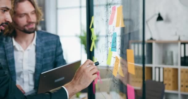 Side view of likable positive successful experienced two bearded coworkers which analyzing opportiunities of business start up project using records on glass wall and sticky notes — Stock Video