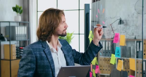 Likable confident skilled successful bearded man standing near glass wall and doing records on it with black marker in modern office room,concept of creativity — Stock Video